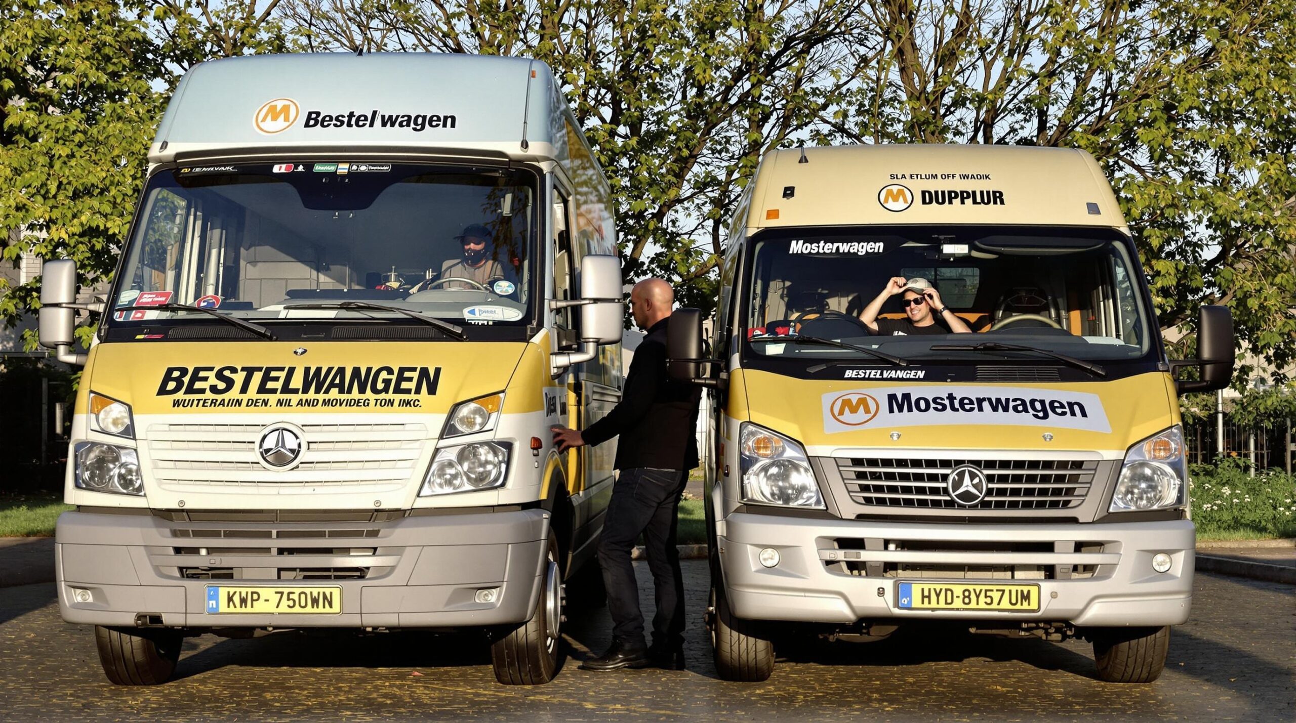 Twee bestelwagens staan naast elkaar geparkeerd op een oprit, met een monteur die onder de motorkap van één kijkt.